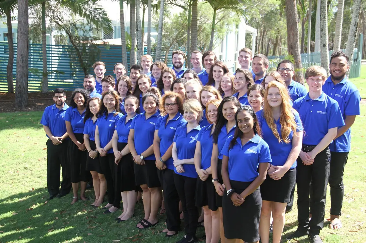 A large NET team in blue shirts standing on a lawn under palm trees