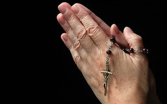Older hands holding a rosary against a black background