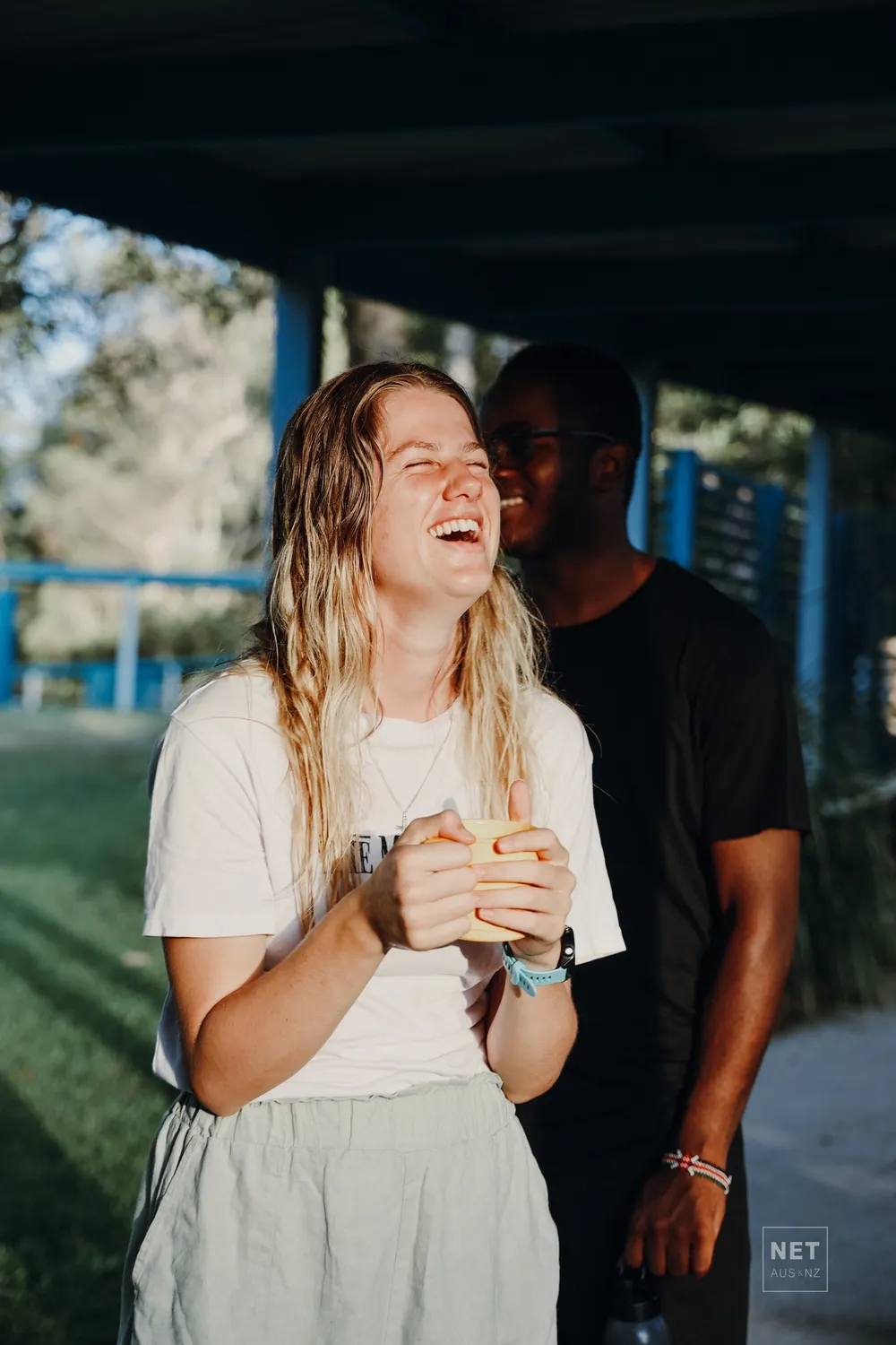 A young woman laughing outdoors, with a young man behind her