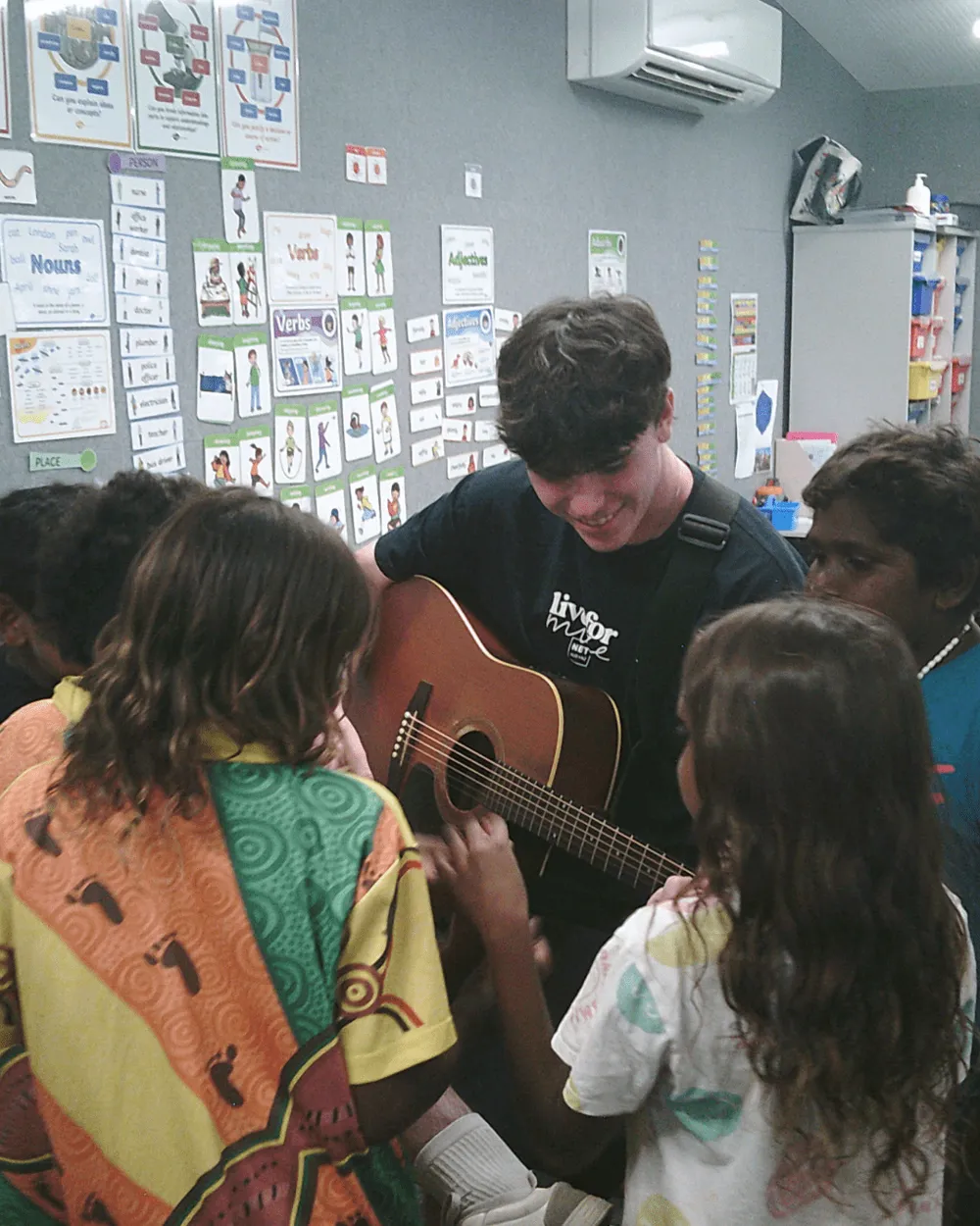 A young man with a guitar among children in a classroom